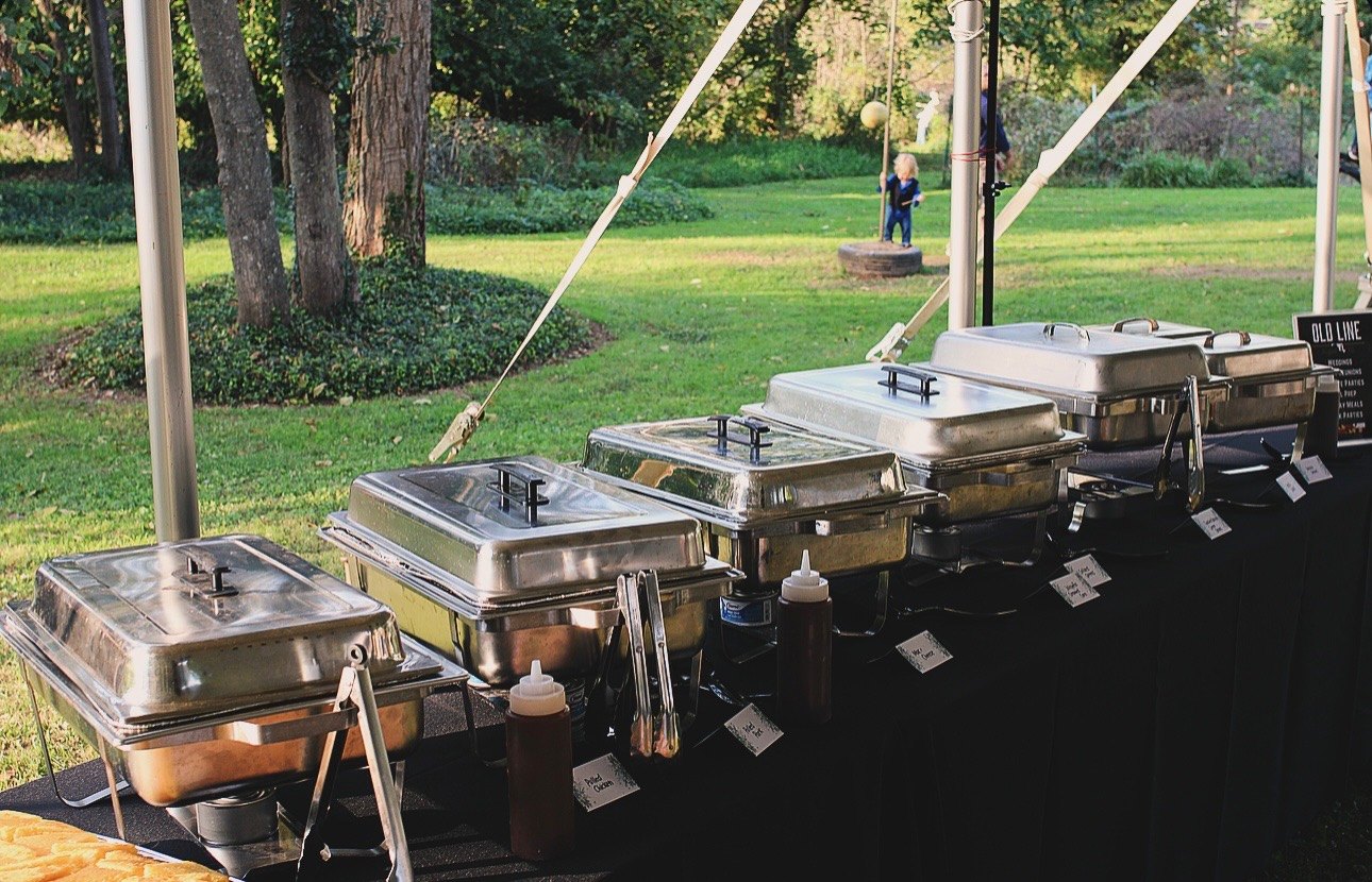 A line of chafing dishes set under a tent on the lawn at a catered event