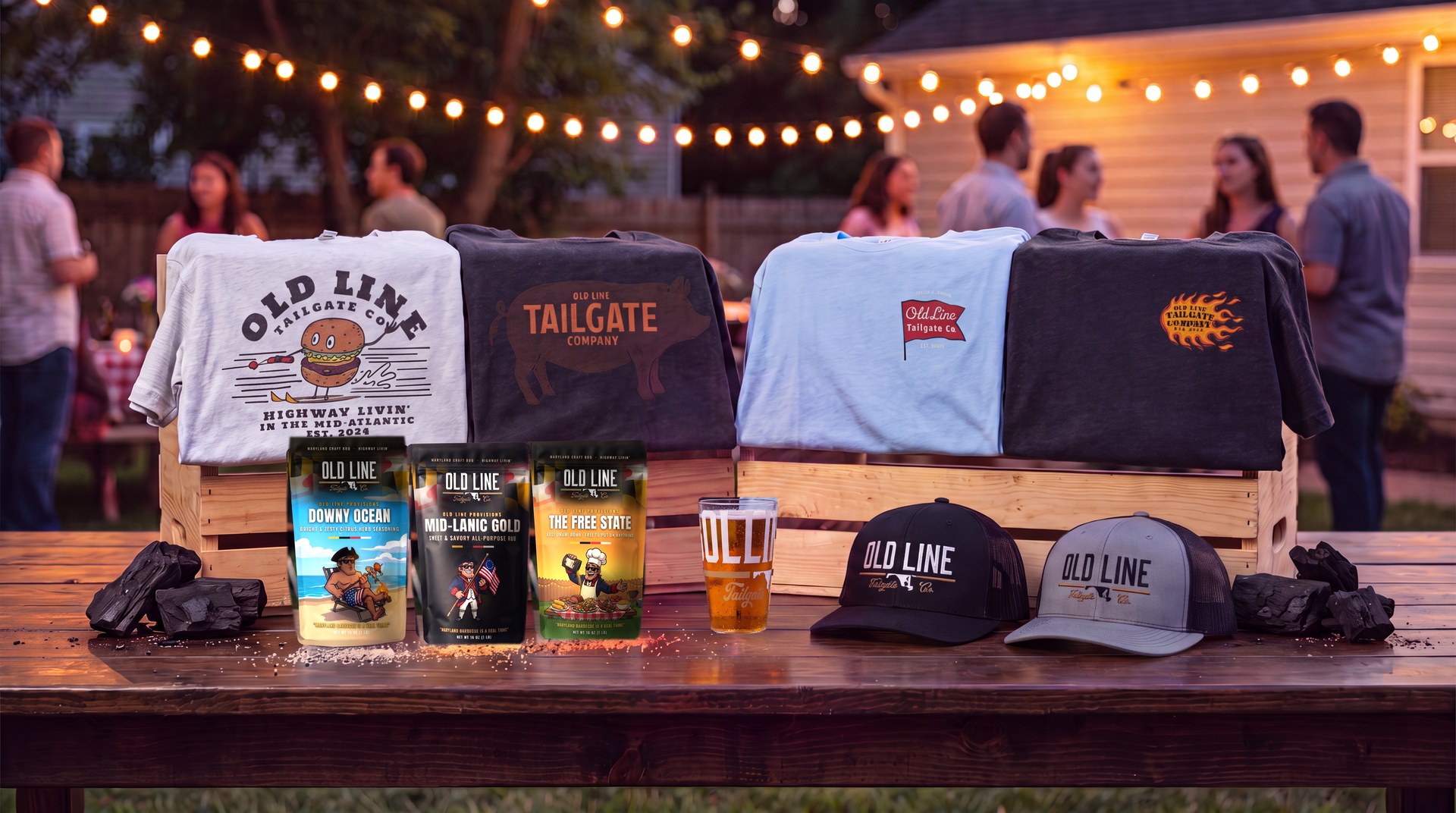 The Old Line Tailgate Co. lineup — tees, trucker hats, Old Line Provisions rubs, charcoal, and a pint glass staged on wooden crates under string lights at a backyard cookout