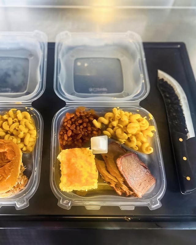 Two carryout boxes from the window: a pulled pork sandwich on the branded bun, and a brisket plate with beans, mac and cheese and cornbread