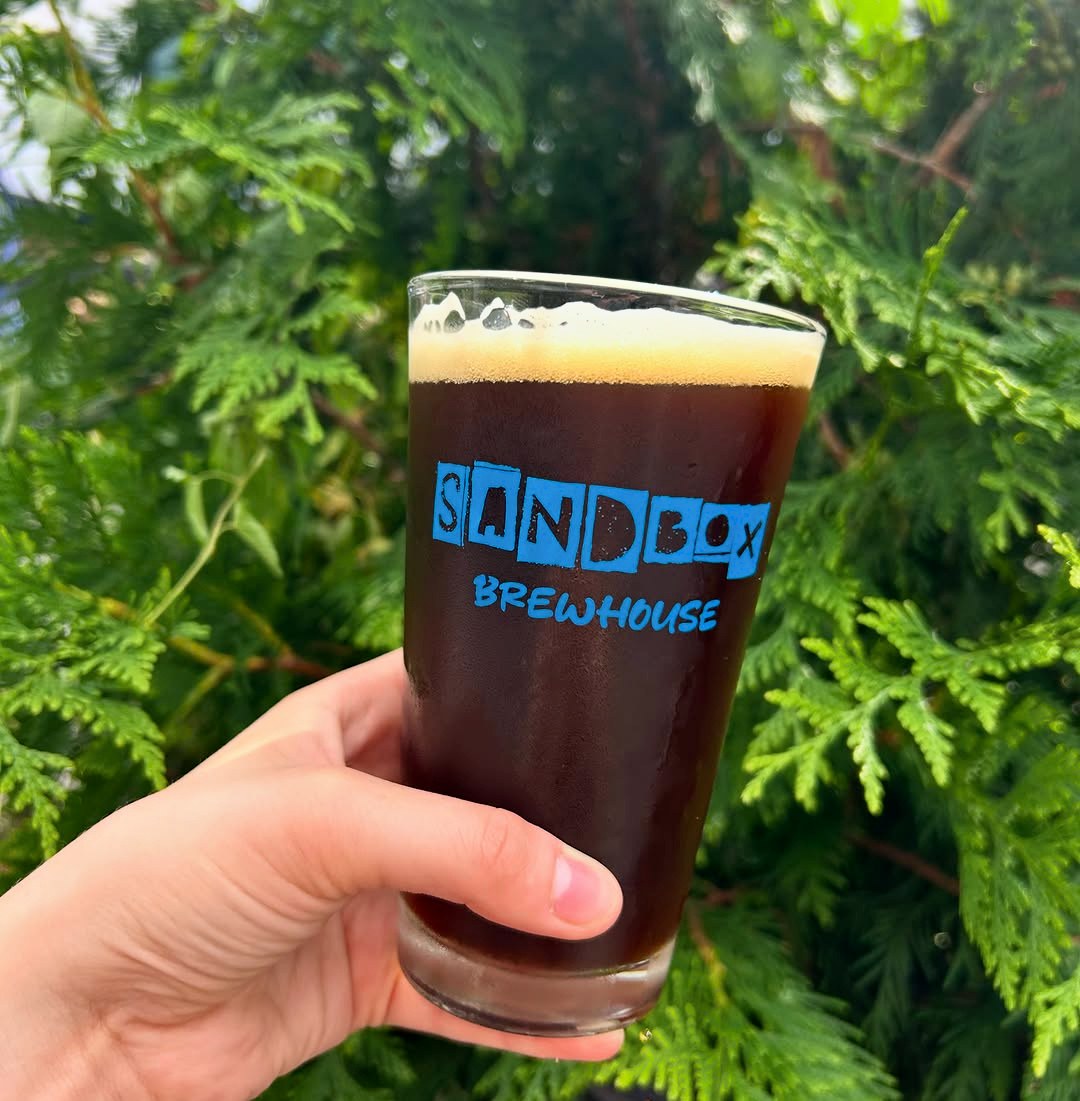 A dark beer in a Sandbox Brewhouse pint glass held up against evergreen branches