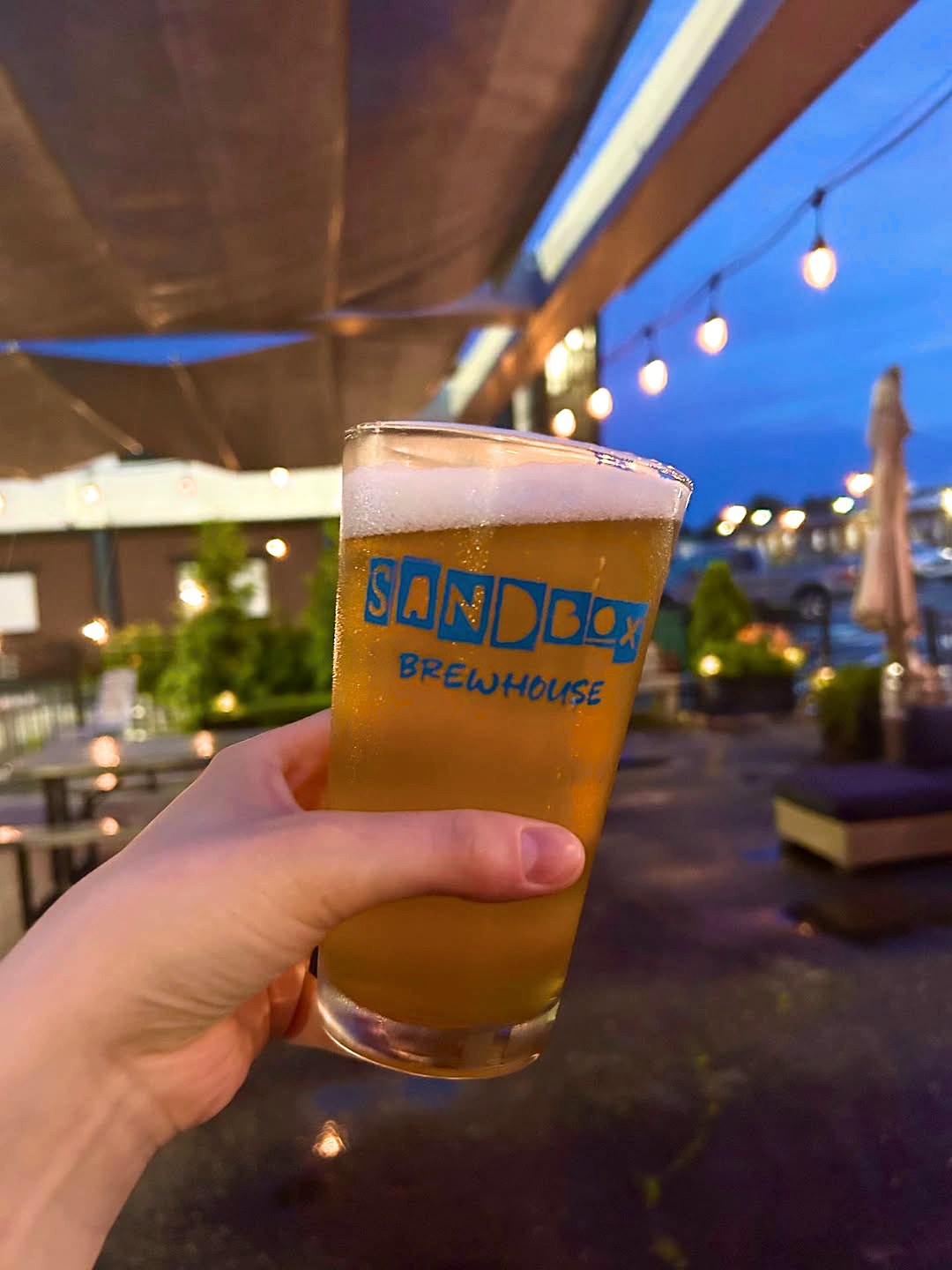 A pint in a Sandbox Brewhouse glass held up on the patio at dusk, string lights glowing overhead