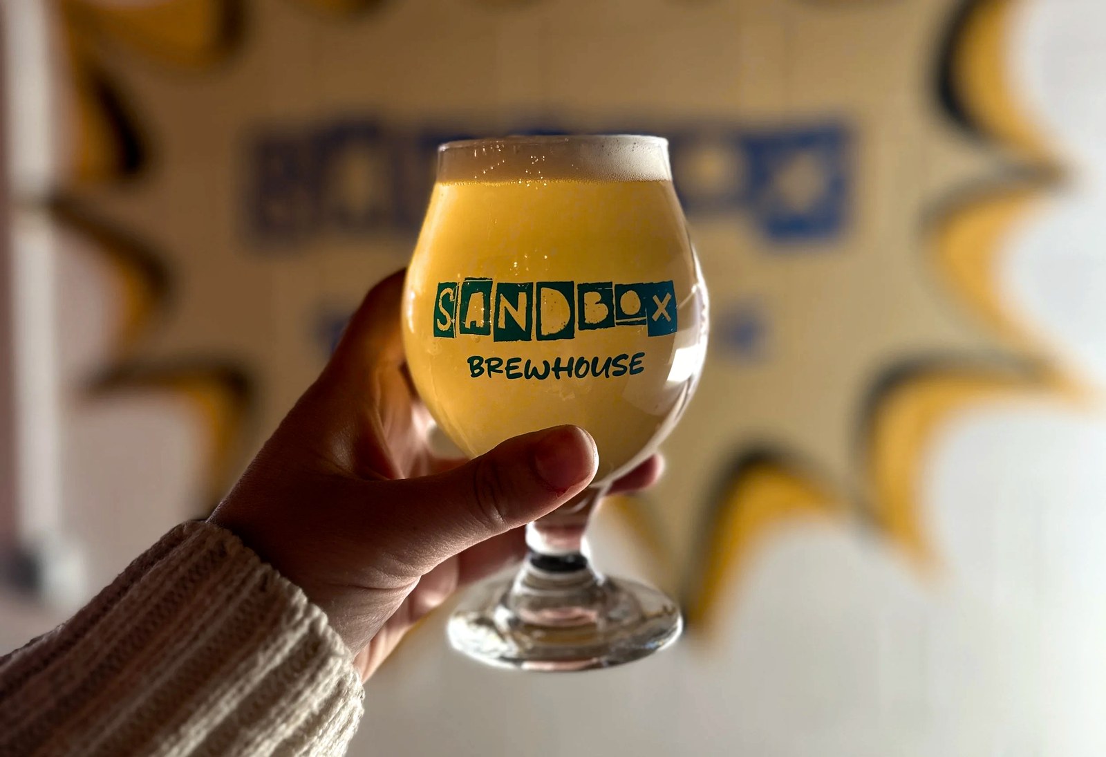 A tulip glass of Sandbox beer held up in front of the Sandbox Brewhouse starburst painted on the taproom wall